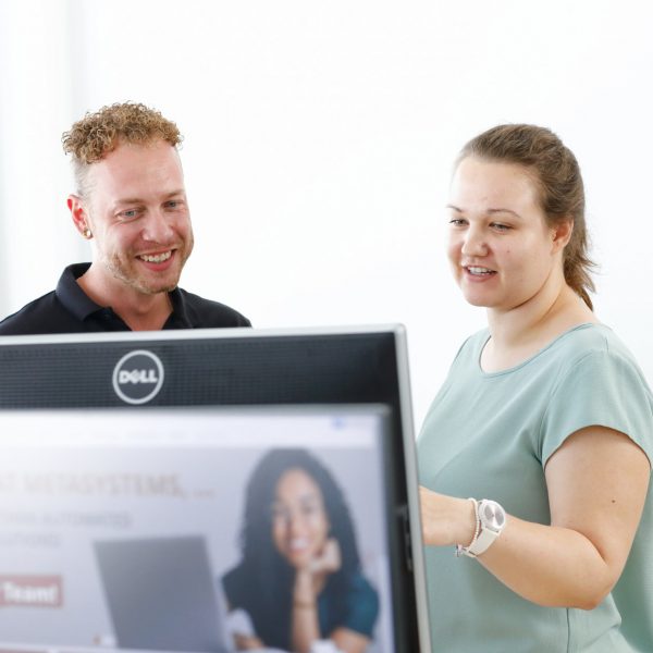 Two colleagues collaborating in front of a computer screen in a bright office, smiling and engaged in discussion.