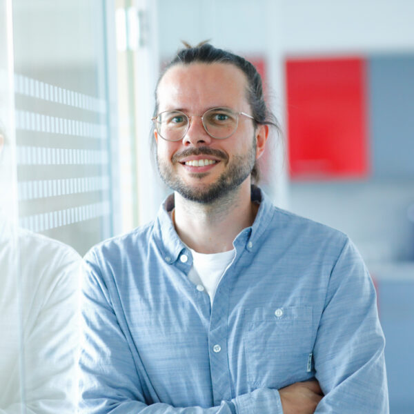 Smiling man with glasses and a beard, wearing a light blue shirt, standing with arms crossed in a bright, modern office environment.