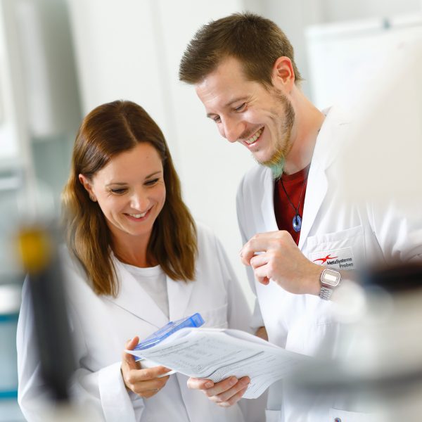 Two scientists in white lab coats smiling and reviewing documents together in a laboratory setting, with visible MetaSystems Probes branding on one coat.