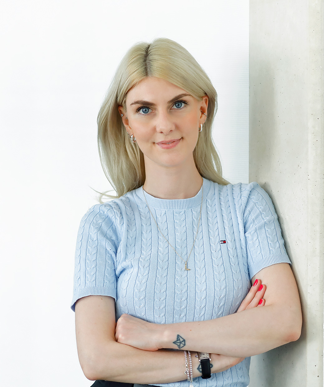 Blonde woman in a light blue top and black trousers, leaning against a pillar with arms crossed, smiling at the camera.