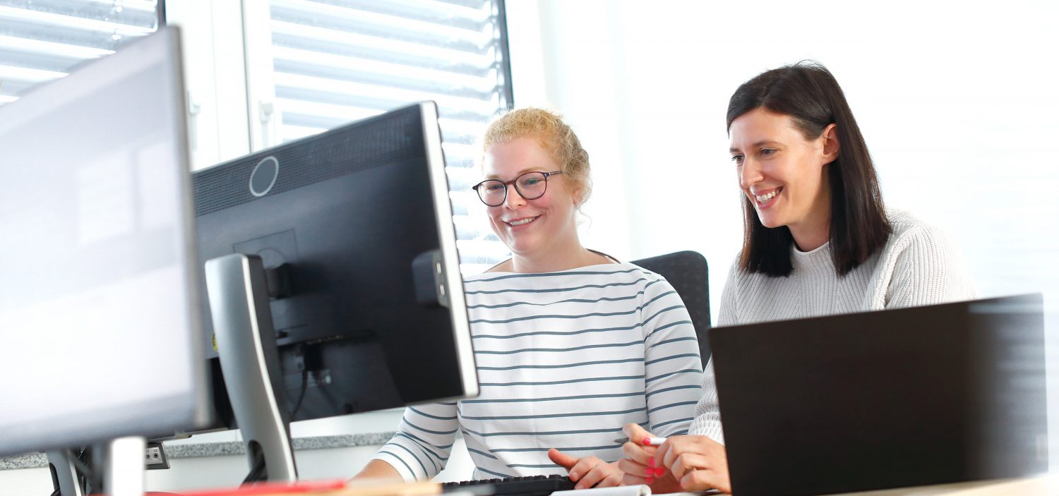 Two colleagues smiling while working together at a desk with laptops and a monitor.