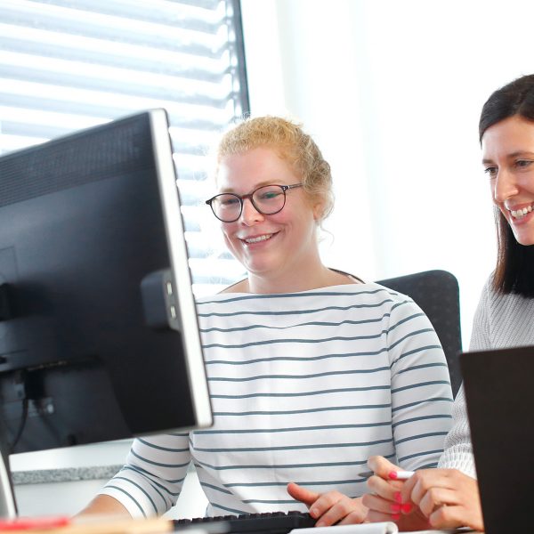 Two colleagues smiling while working together at a desk with laptops and a monitor.